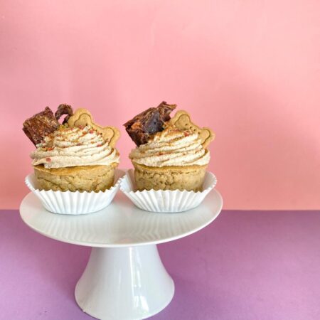 2 cupcakes para perro con ingredientes naturales decorados con crema pastelera apta para perros, snack deshidratado y galleta para perro, sobre fondo rosa.